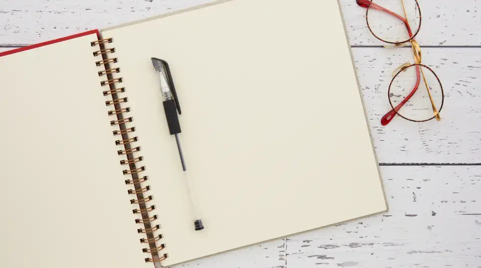 An open spiral notebook with a pen, and a pair of glasses, on white-painted wooden boards.