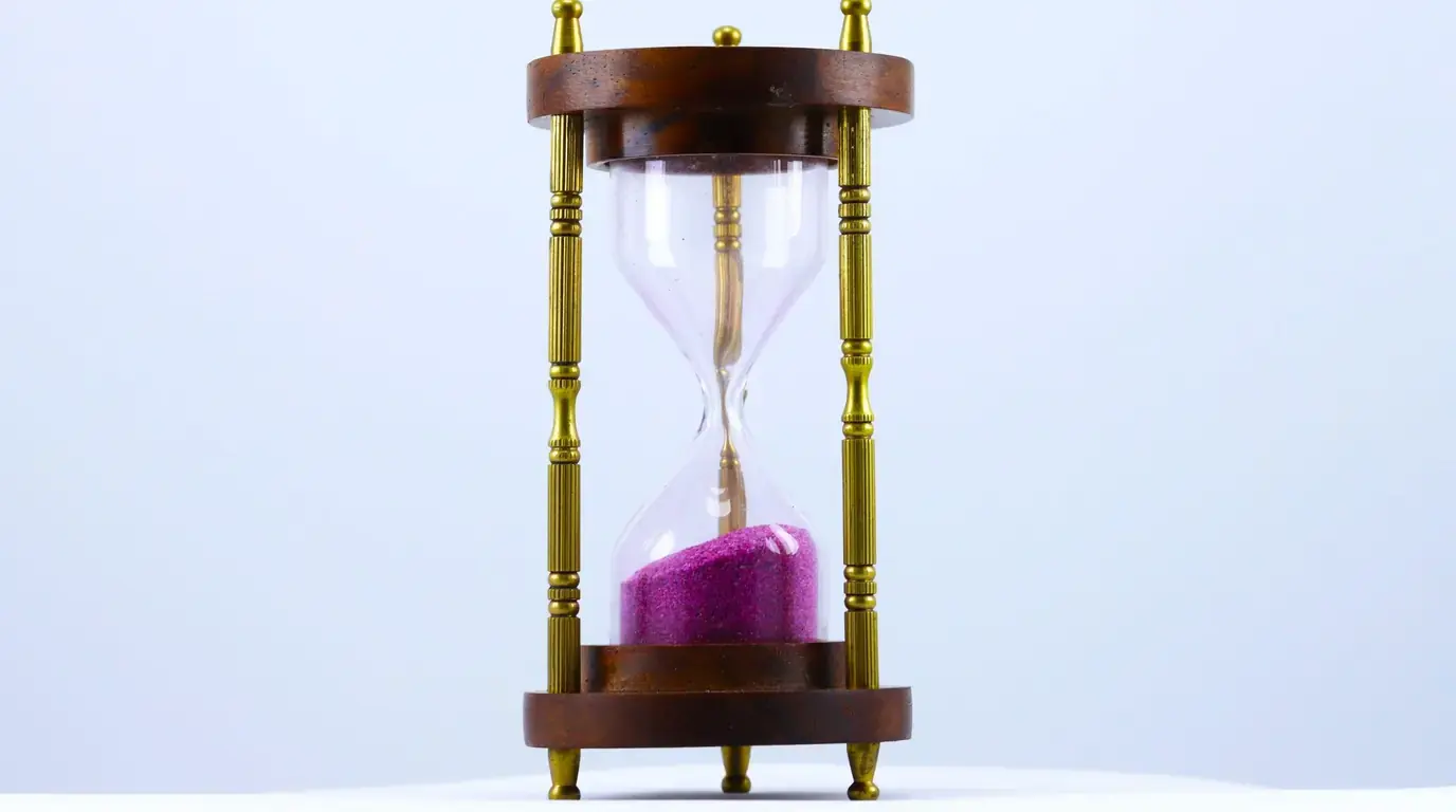 A brass and wood hourglass with pink sand against a pale blue background.