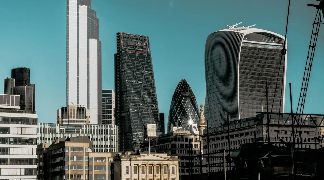 Glass office towers in varied shapes, wedge, curve and latticed dome, rising over older stone rooftops beneath a deep teal sky.