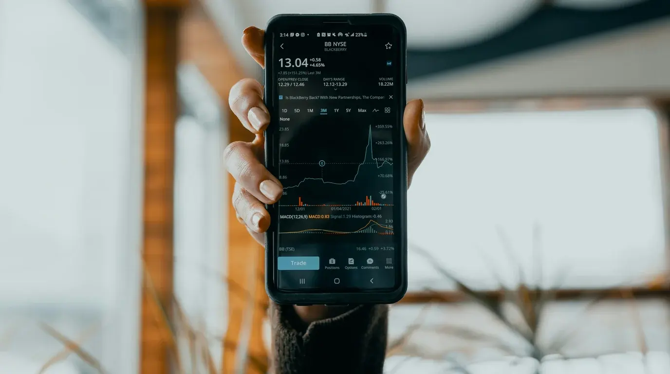 A hand holding up a phone displaying a dark stock-tracking app with a price chart, volume bars and indicator lines.