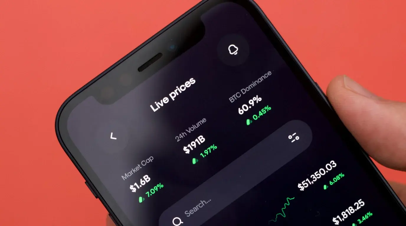 Close-up of a phone tilted against a coral-red background, its dark screen listing live market prices and percentage changes.