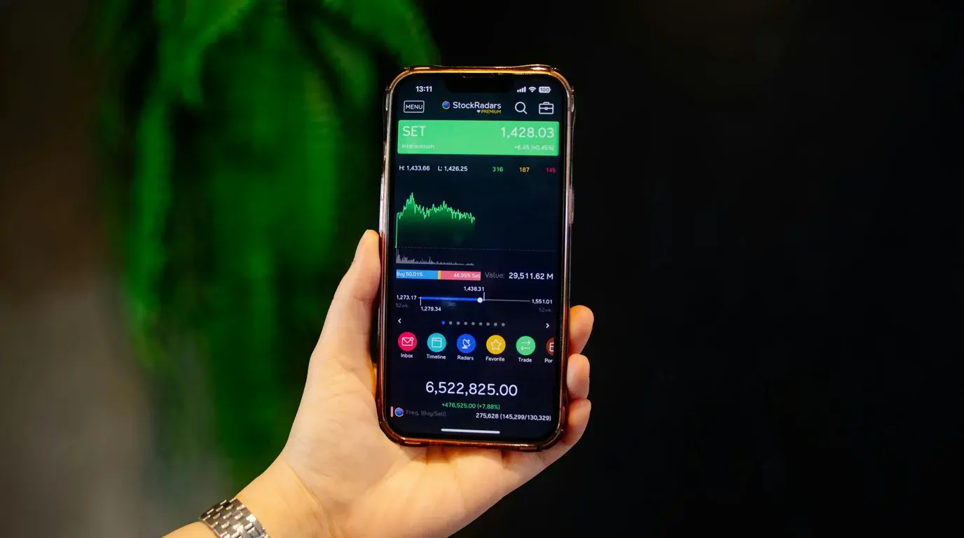 A hand holding a phone showing a green market index panel and charts, a blurred green plant and dark background behind.