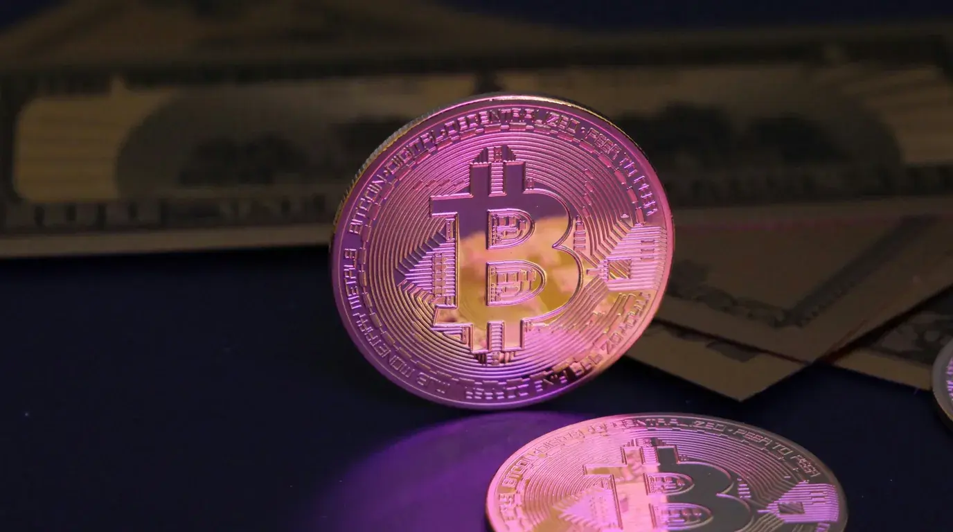 A gold-coloured Bitcoin token standing on end among others lying flat, banknotes behind, lit pink and purple.