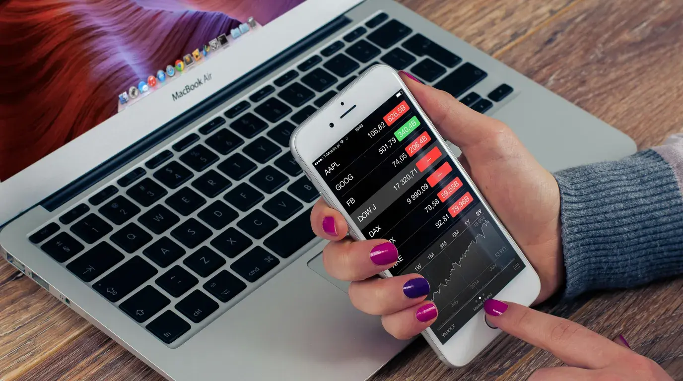 Brightly manicured hands holding a phone with a red and green market ticker list, resting over a laptop keyboard on wood.