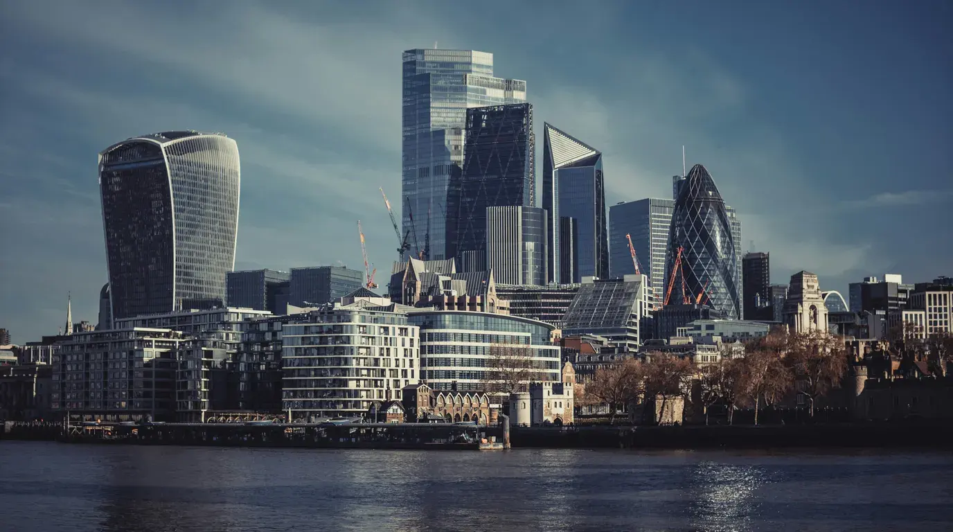 Tall glass towers and cranes above riverside apartment blocks and a stone bastion, seen across dark rippling water.