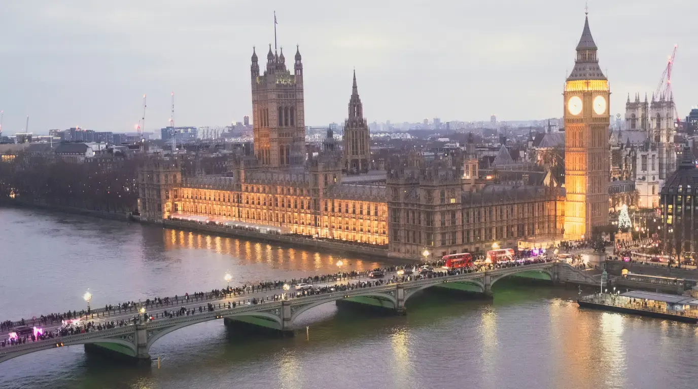 A floodlit Gothic riverside parliament and clock tower at dusk, with crowds crossing the arched bridge below.