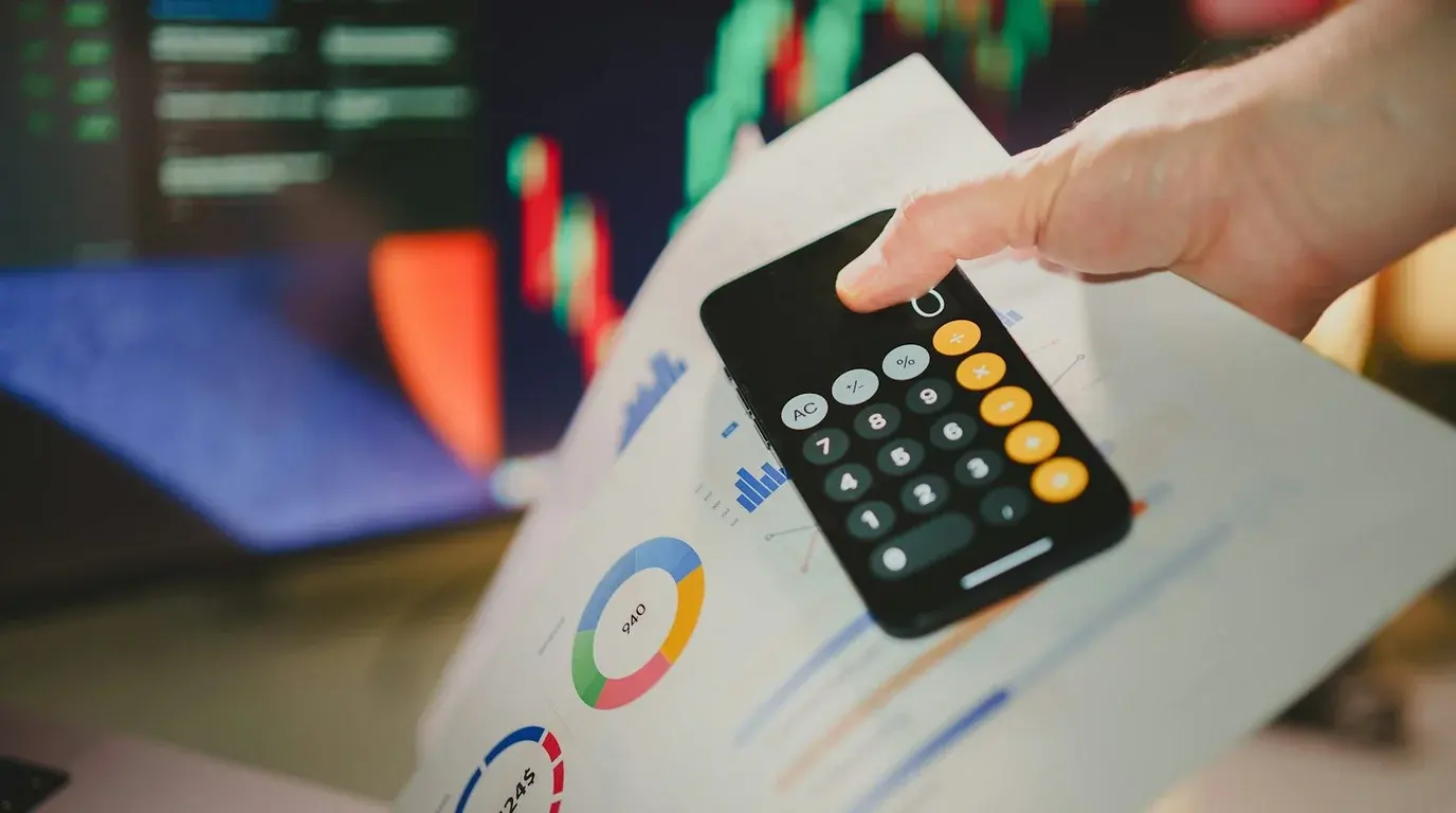 A hand reaching for a phone showing a calculator, resting on printed sheets of pie charts and bar graphs, screens blurred behind.