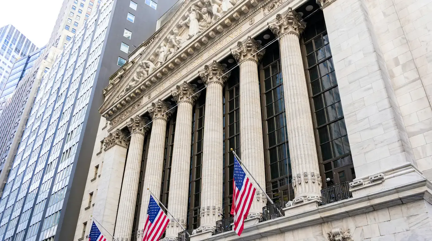Low angle on a neoclassical stone facade of tall fluted columns and a sculpted pediment, with three flags hanging from poles above the street