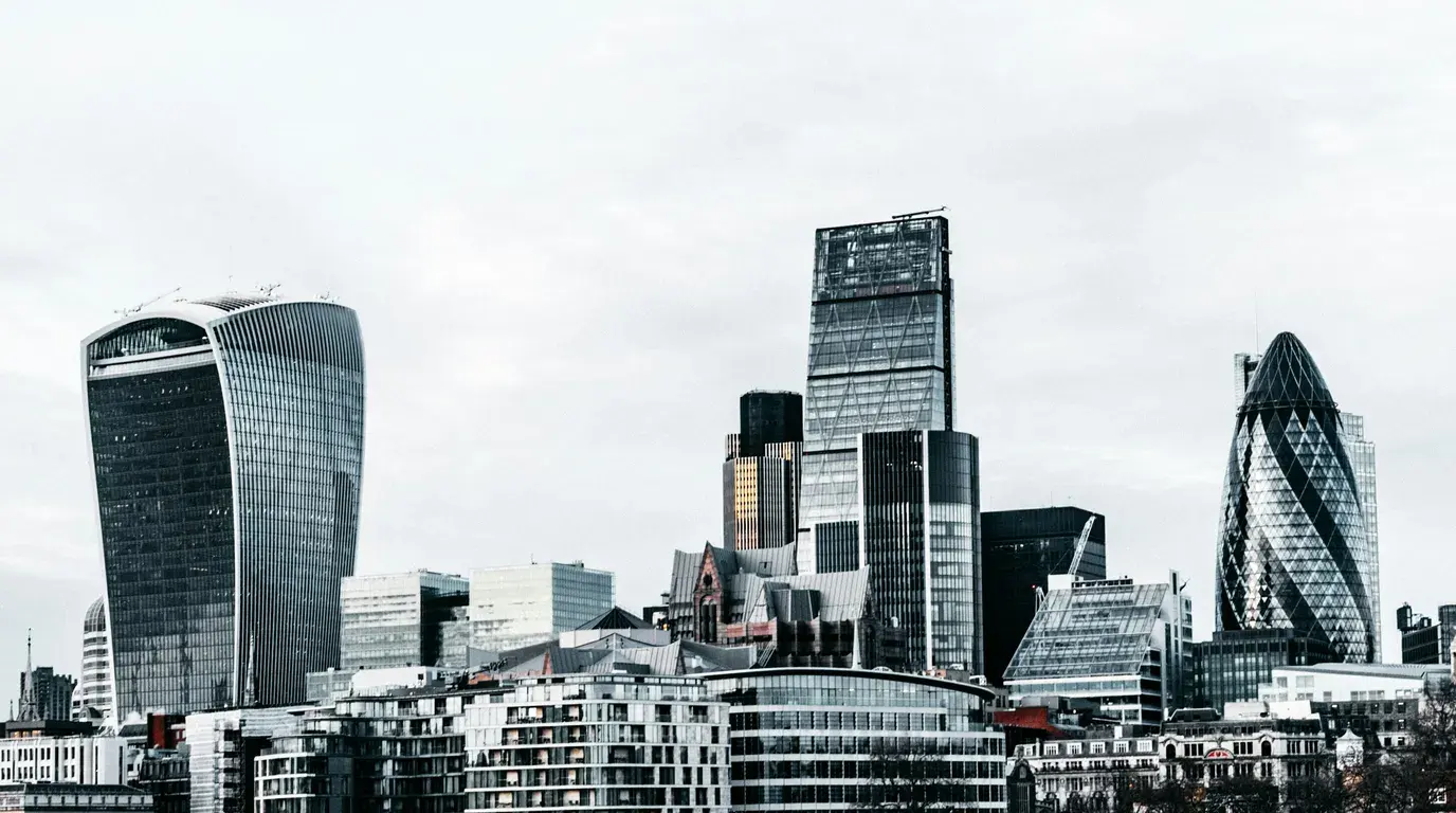 Desaturated skyline of glass and steel office towers in varied shapes, rising over lower brick and concrete blocks under a flat white sky