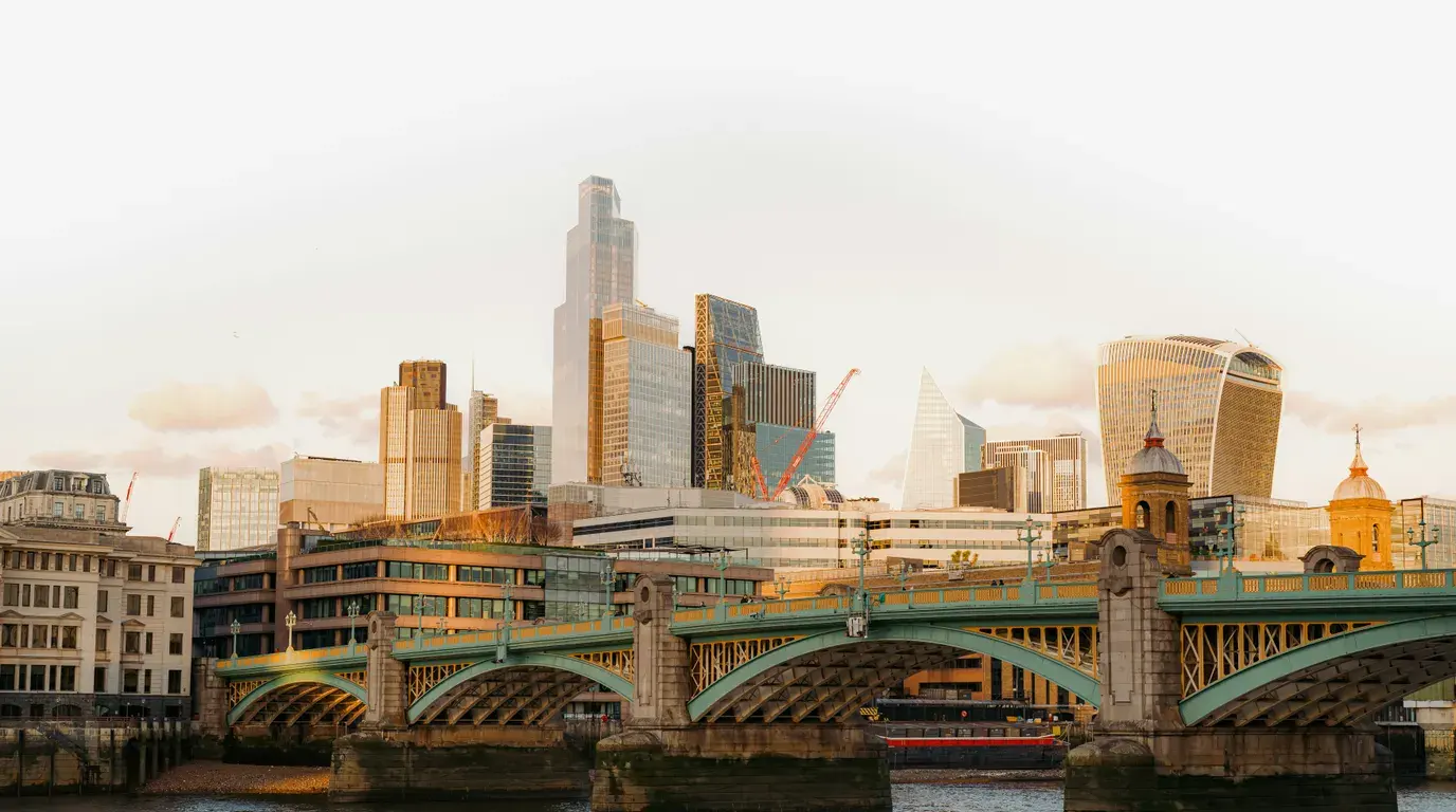 Green cast-iron arched bridge spanning a wide river at golden hour, with clustered glass towers and cranes catching warm light behind it