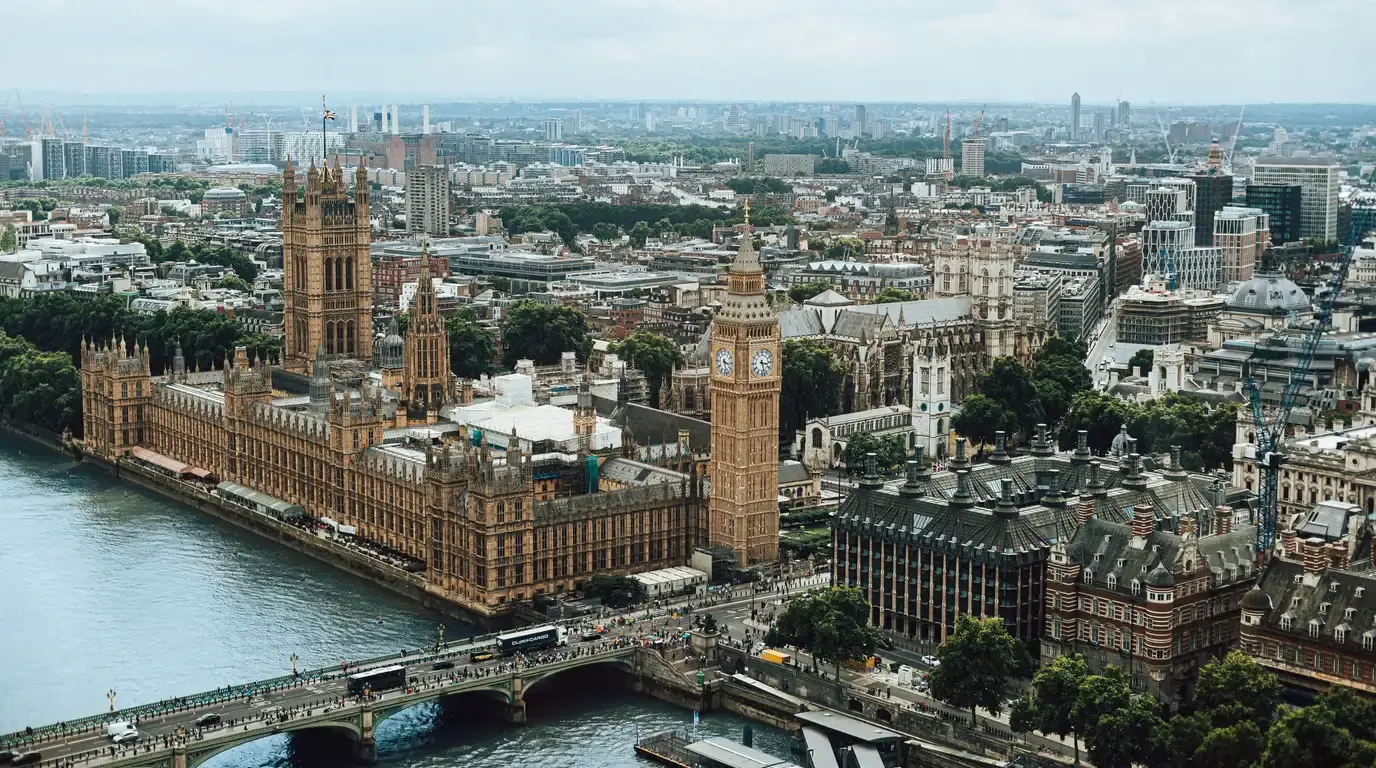Aerial view across a river of a sprawling Gothic stone palace with a tall clock tower, traffic crossing the bridge and rooftops stretching to the horizon