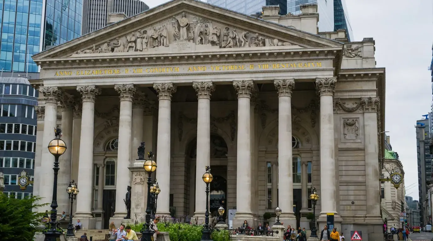 Grand stone building with a columned portico, carved pediment and gilded inscription, ornate black lampposts and people sitting on the steps below