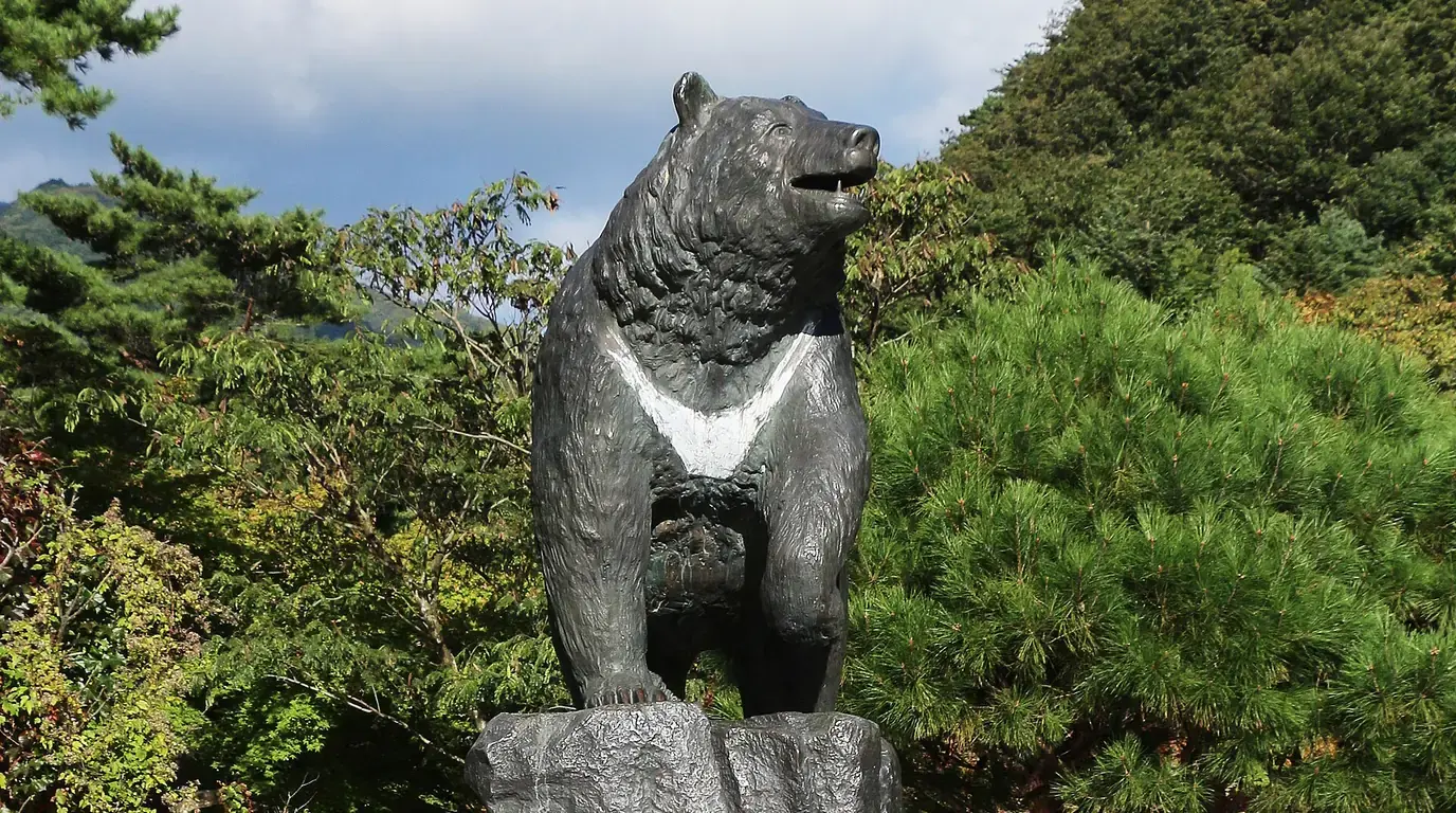 A bronze statue of a bear standing on a rock, surrounded by pine trees under a blue sky.