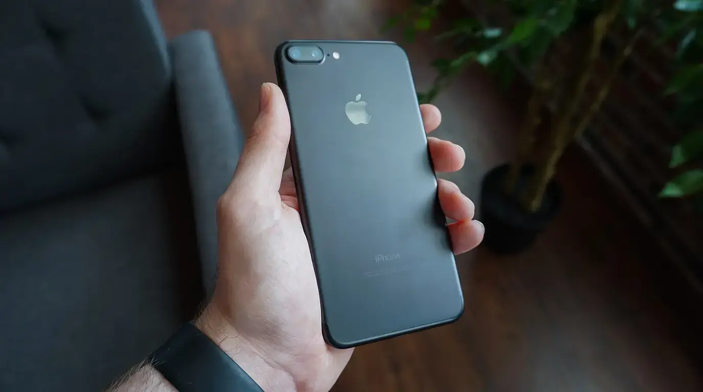 A hand holding a black smartphone with its back facing the camera, in a dimly lit room